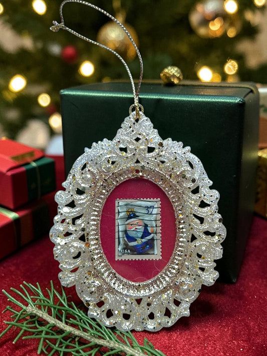 Addobbo natalizio artigianale italiano con Babbo Natale blu su francobollo, cornice ovale dorata tra pacchi regalo