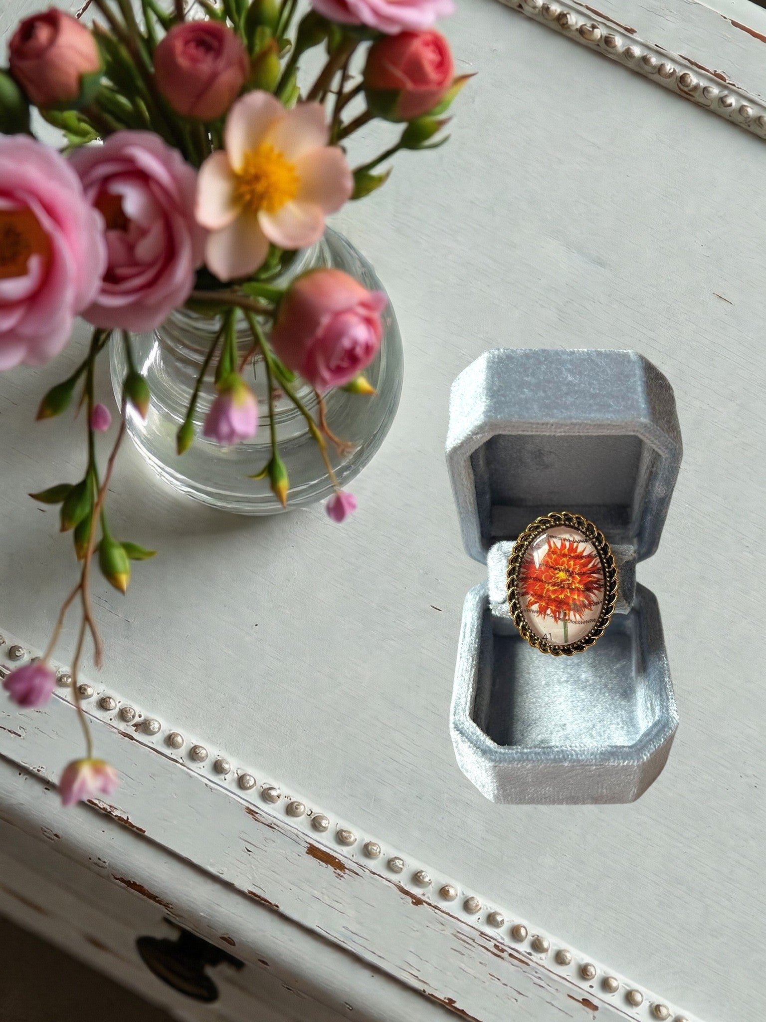 Anello artigianale italiano con dalia rosso-arancio su portanelli azzurro davanti a una finestra, montatura
ovale color oro