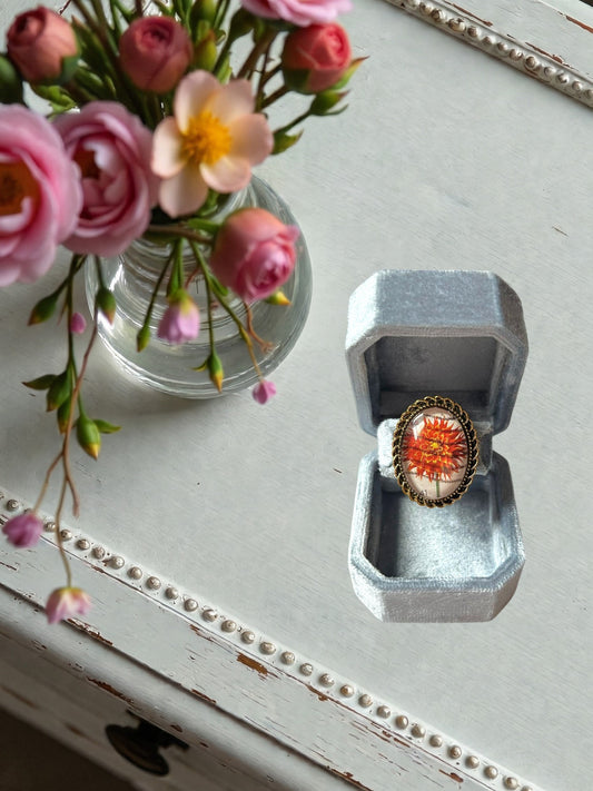 Anello artigianale italiano con dalia rosso-arancio su portanelli azzurro davanti a una finestra, montatura
ovale color oro
