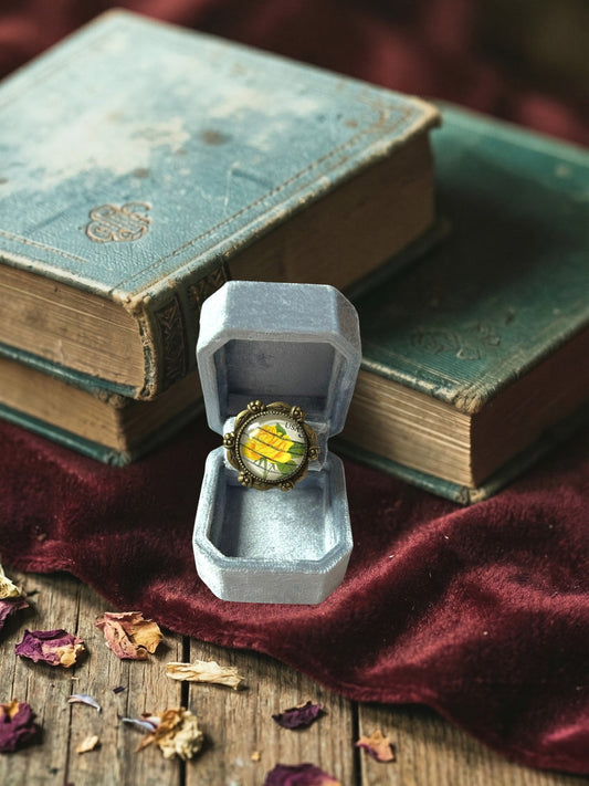 Anello artigianale italiano con rosa gialla su portanelli azzurro davanti a una finestra, montatura bronzo
a roselline