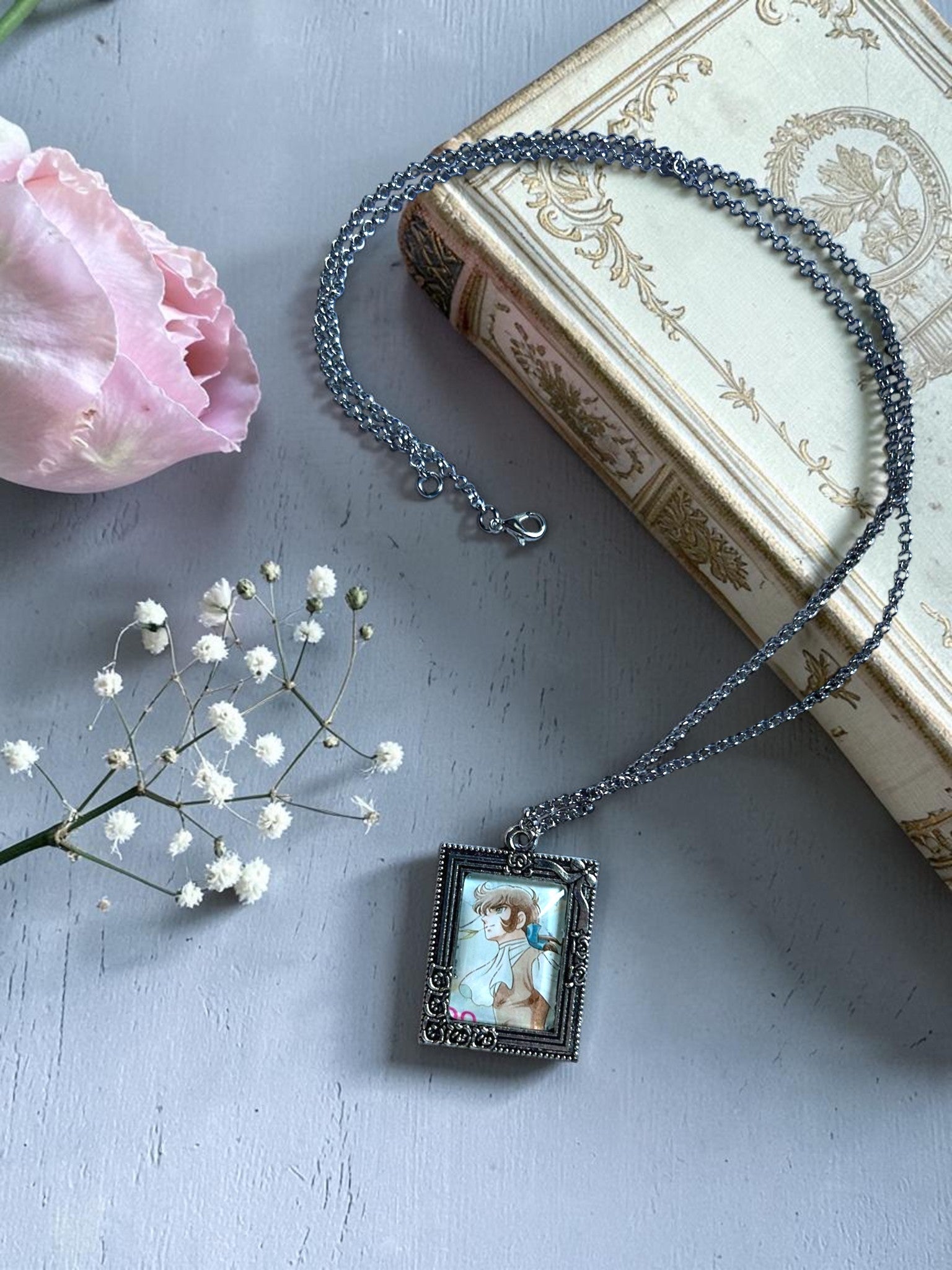 Collana con ciondolo rettangolare argentato e francobollo d'epoca di André, personaggio di Lady Oscar, su libro antico