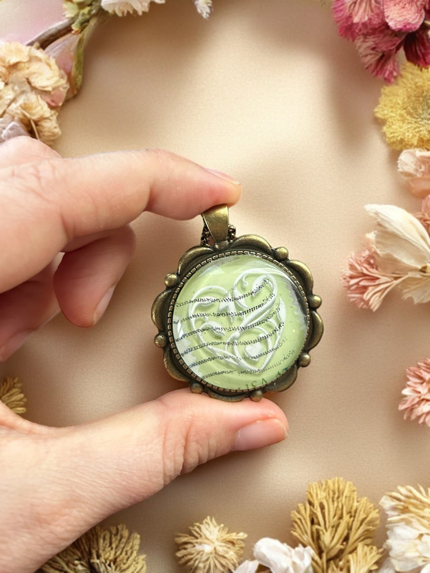 Collana con ciondolo tondo color bronzo e francobollo con cuore bianco su verde, indossata tra fiori secchi, artigianato, Italia