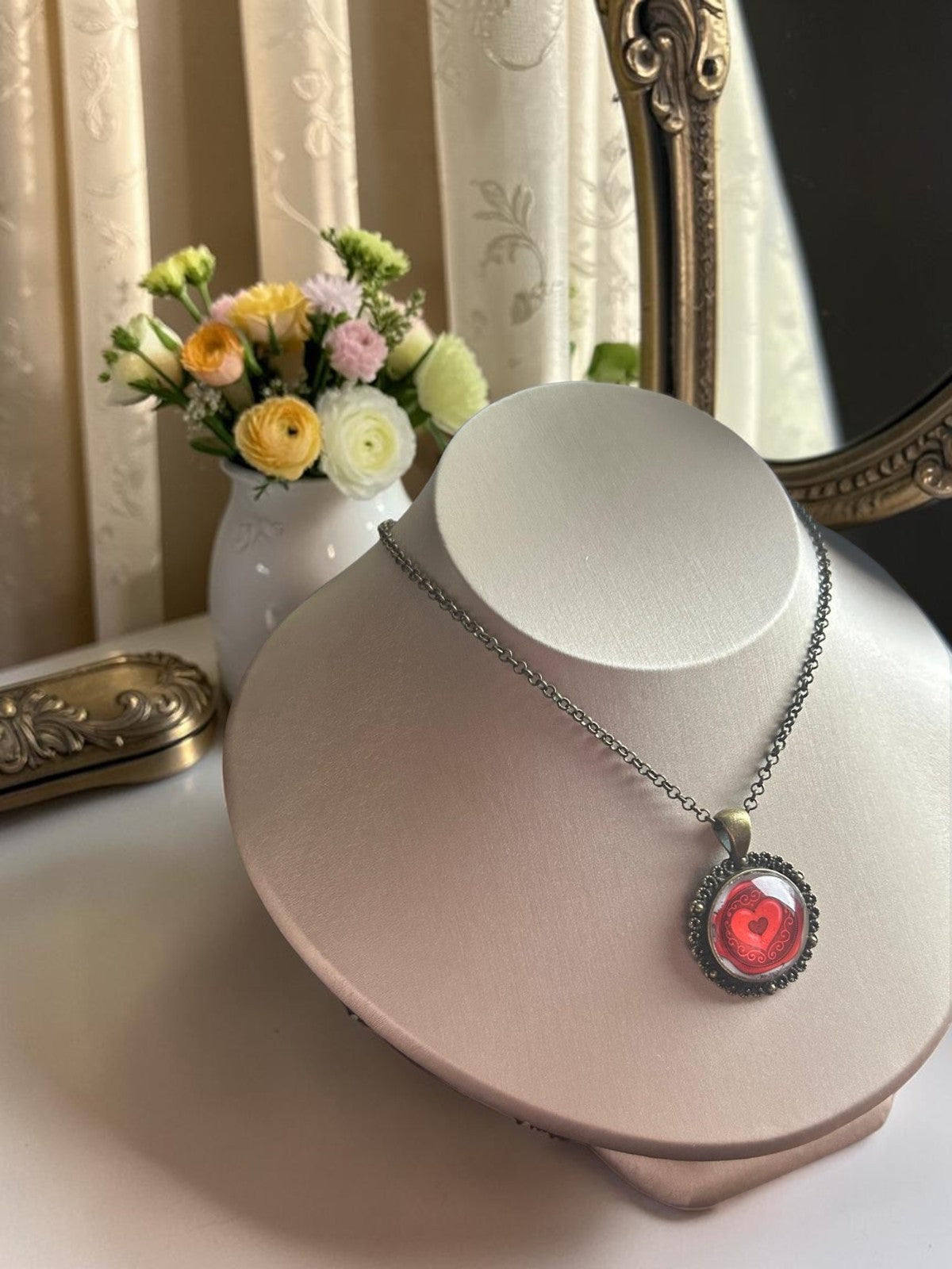 Collana mignon con ciondolo tondo e cuore rosso, esposta su busto tra fiori e specchio dorato