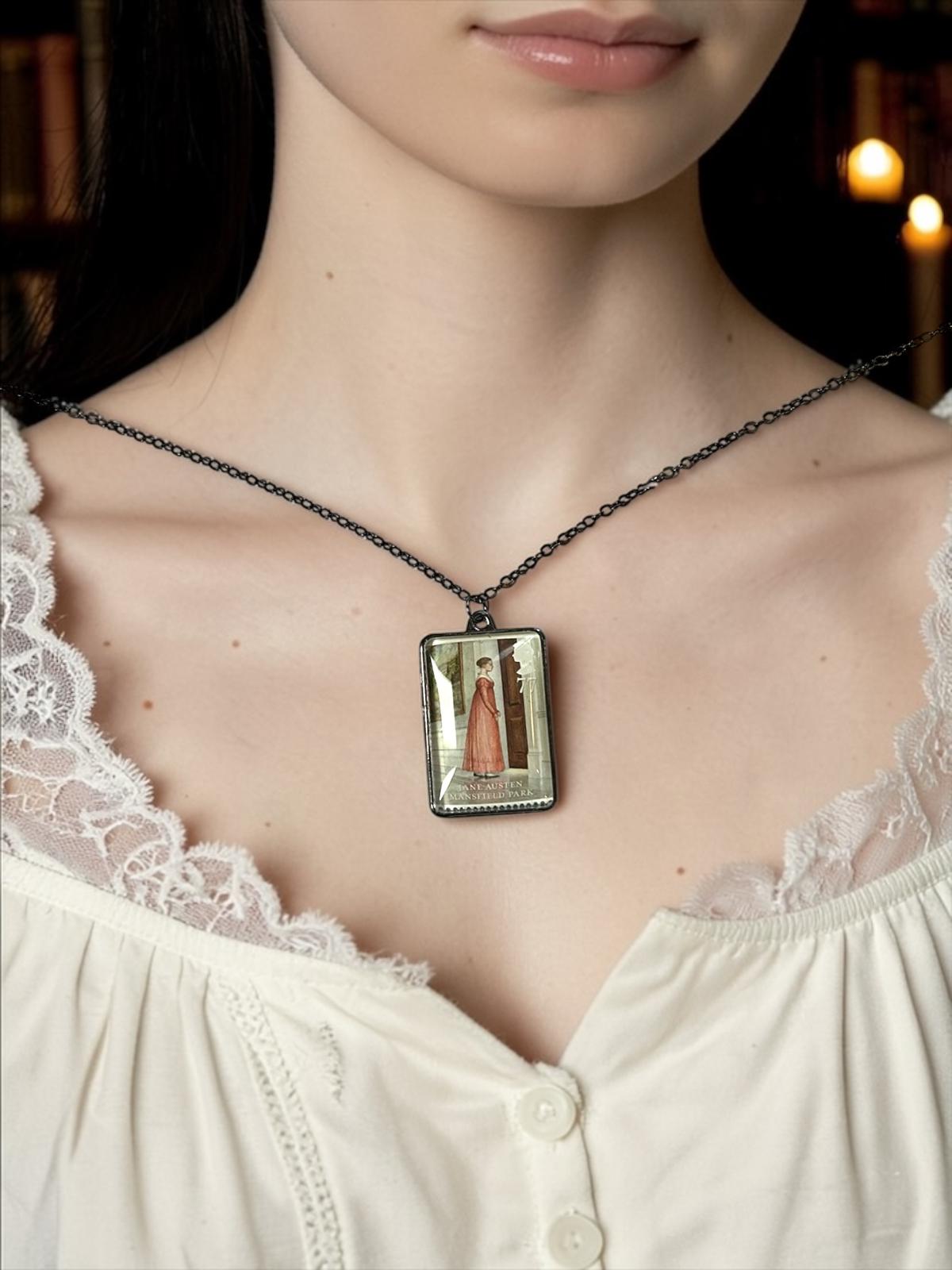 Collana Jane Austen con il francobollo Royal Mail del 2013 dedicato a Mansfield Park, illustrato da Angela Barrett, incastonato in una montatura rettangolare di metallo scuro. La collana è indossata su un busto.