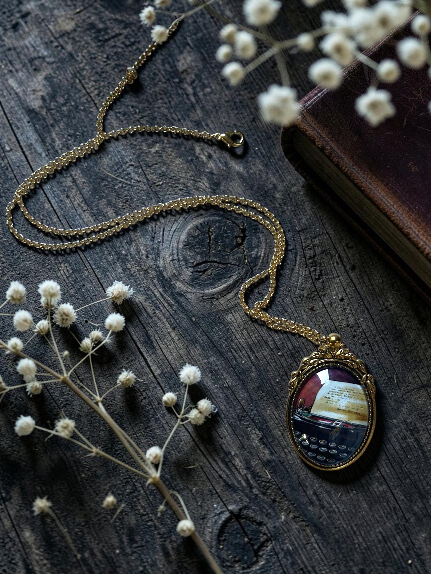 Collana dorata con ciondolo ovale e macchina da scrivere di Agatha Christie, distesa tra fiori bianchi e un libro