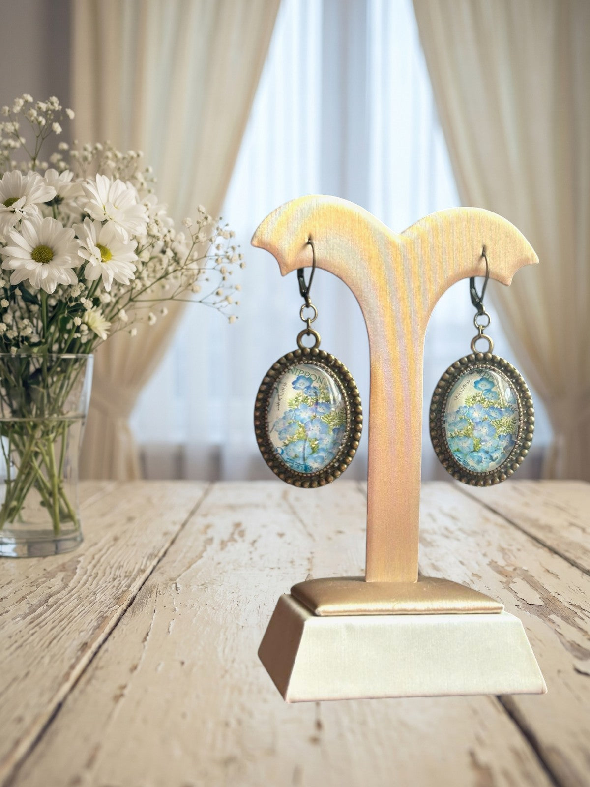 Orecchini artigianali italiani con fiori azzurri nemofila su portaorecchini beige-oro davanti a una finestra