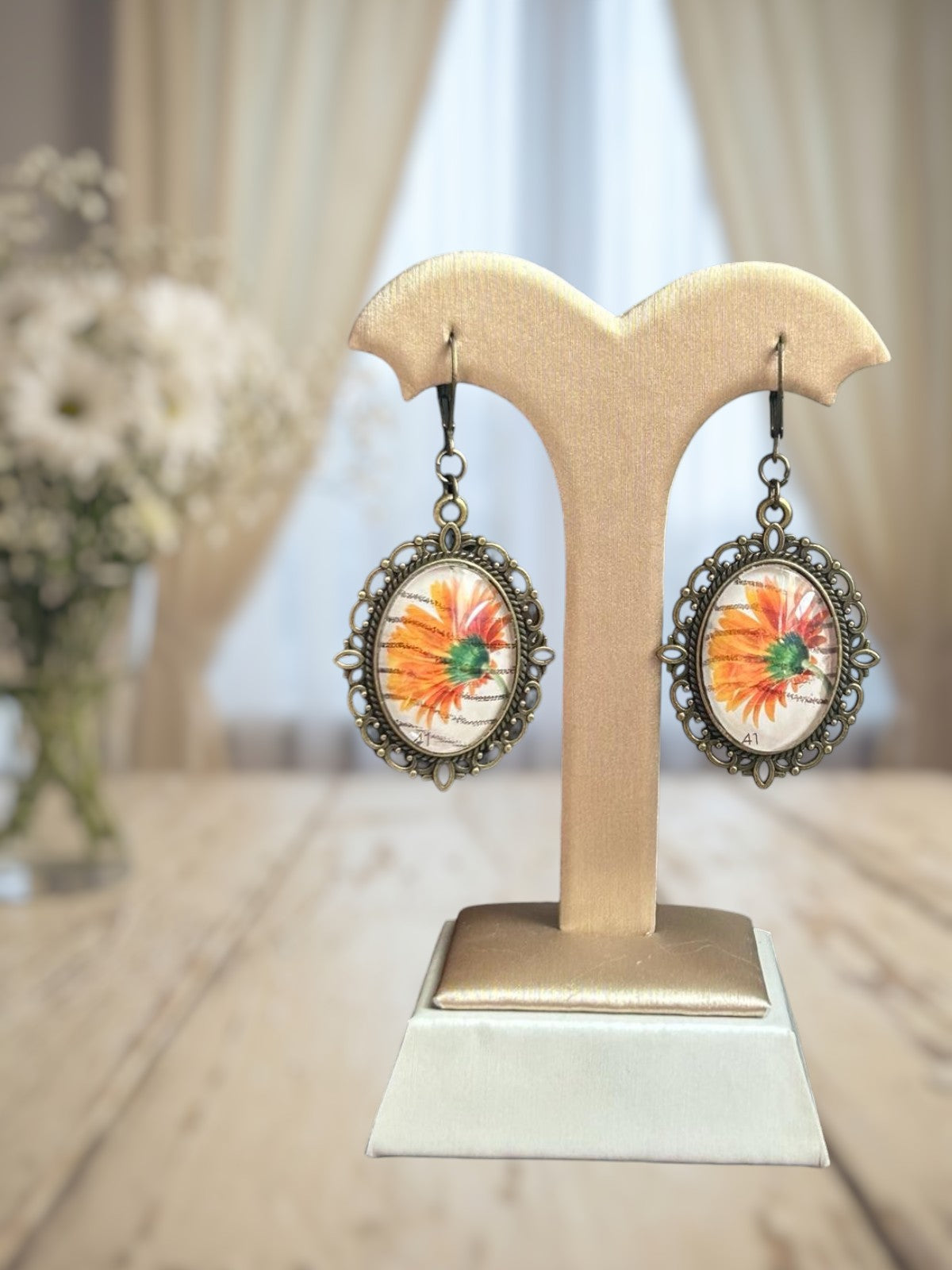Orecchini artigianali italiani con gerbera arancione su portaorecchini beige-oro davanti a una finestra