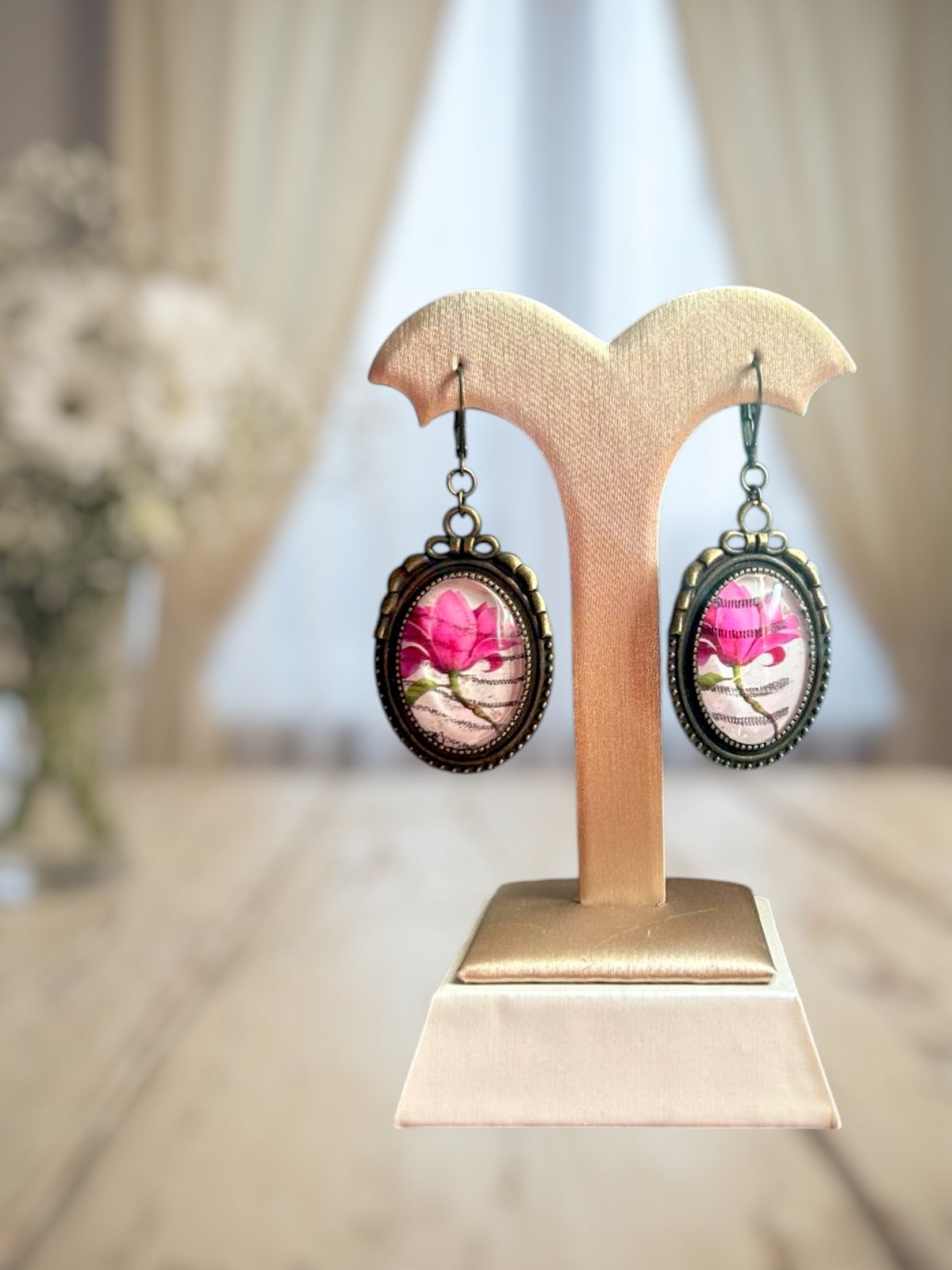 Orecchini artigianali italiani con magnolia rosa su portaorecchini beige-oro davanti a una finestra