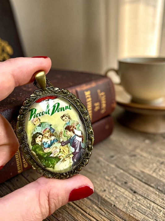 Collana artigianale italiana tenuta in mano, ciondolo ovale con la copertina vintage di Piccole Donne, libri sullo sfondo
