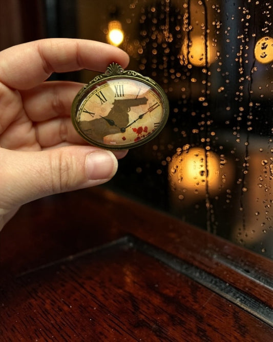 Spilla ovale Lady Nostalgia con francobollo di Agatha Christie: quadrante d'orologio, sagoma di rivoltella e gocce di sangue.