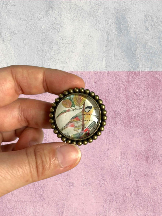 Spilla artigianale con francobollo malese di uccellini dal petto arancio, in mano, sfondo rosa, Italia